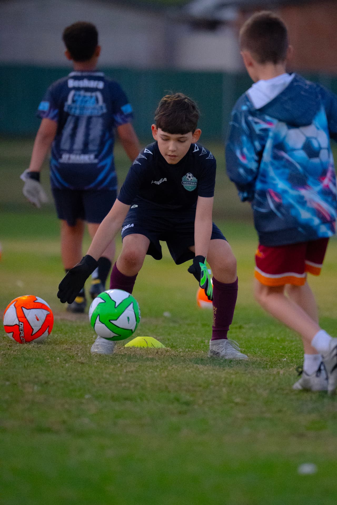 NextGen goalkeeper in a black kit reaching low for the ball in a ground-catch drill