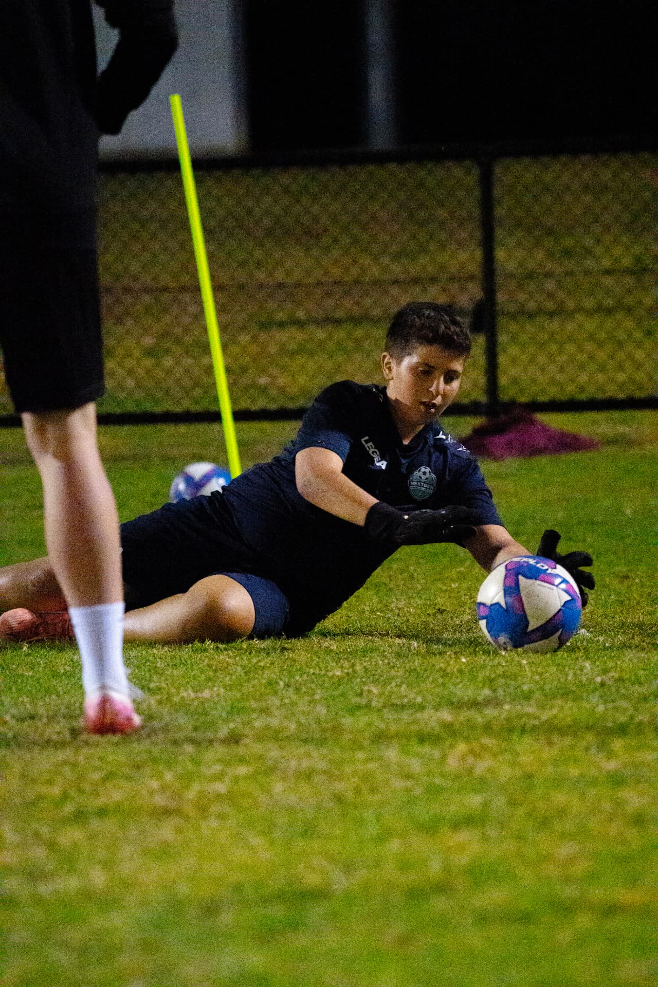 NextGen goalkeeper diving low to save a shot during an evening session